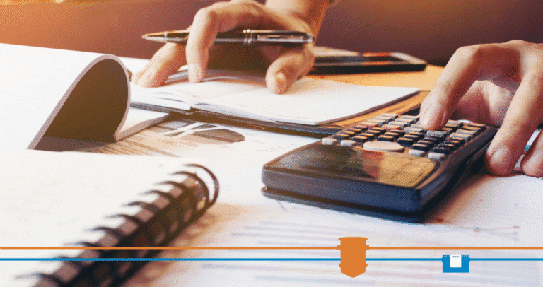 A person uses a calculator and takes notes on paper at a desk covered with documents, notebooks, and a pen, possibly reviewing costs for an automatic grease removal system or managing financial records for a self-cleaning grease trap.