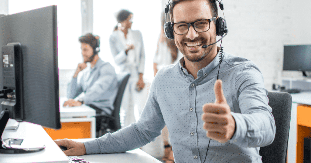 A man wearing glasses and a headset sits at a desk in an office, smiling and giving a thumbs-up gesture—perhaps celebrating the installation of an automatic oil and grease separator. Other people are working at desks in the background.