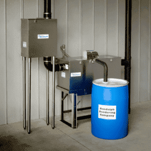 An In-line Strainer with pipes and an automatic grease removal system connects to a blue Randolph Rendering Company barrel, set in a clean industrial space with a gray paneled wall.
